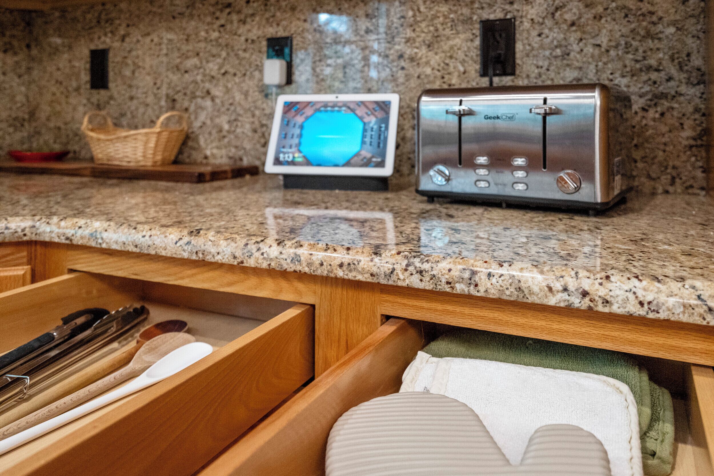A kitchen counter with a toaster, a digital screen, and a wooden drawer with utensils.