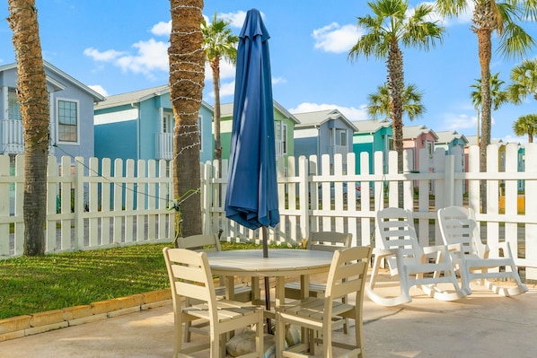 A patio with a round table and chairs, a blue umbrella, and a white picket fence. - Beachside Comfort | 2BR Condo Near Pier Park + Daily Freebies! (Panama City Beach)