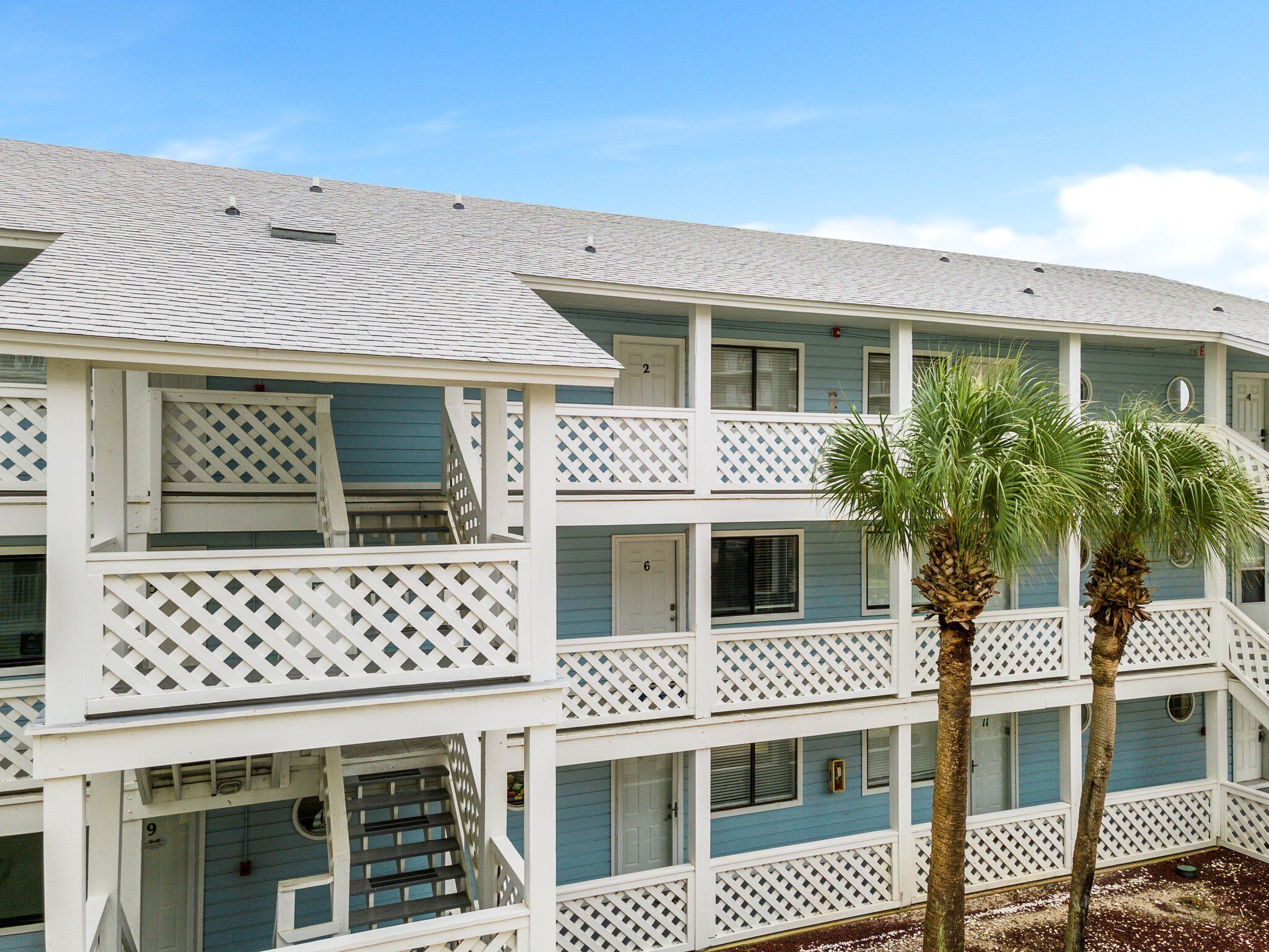 A multi-story hotel with a white exterior and blue accents, featuring balconies and a palm tree in the foreground.