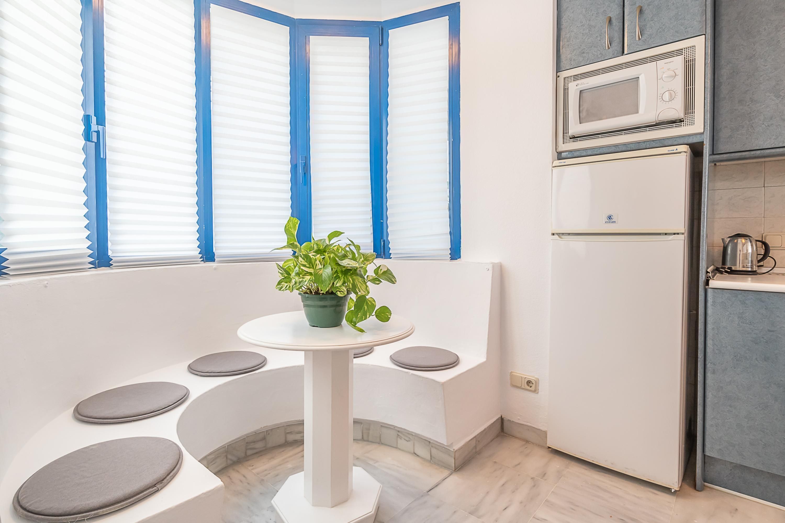 Une cuisine moderne dotée d’un four à micro-ondes et d’un réfrigérateur encastrés, d’un banc blanc avec des assises rembourrées et d’une plante en pot posée sur une petite table.