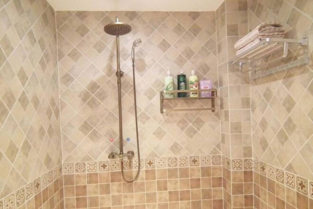 Zona de ducha con cabezal de ducha de lluvia, un cabezal de ducha de mano y un estante con artículos de aseo.