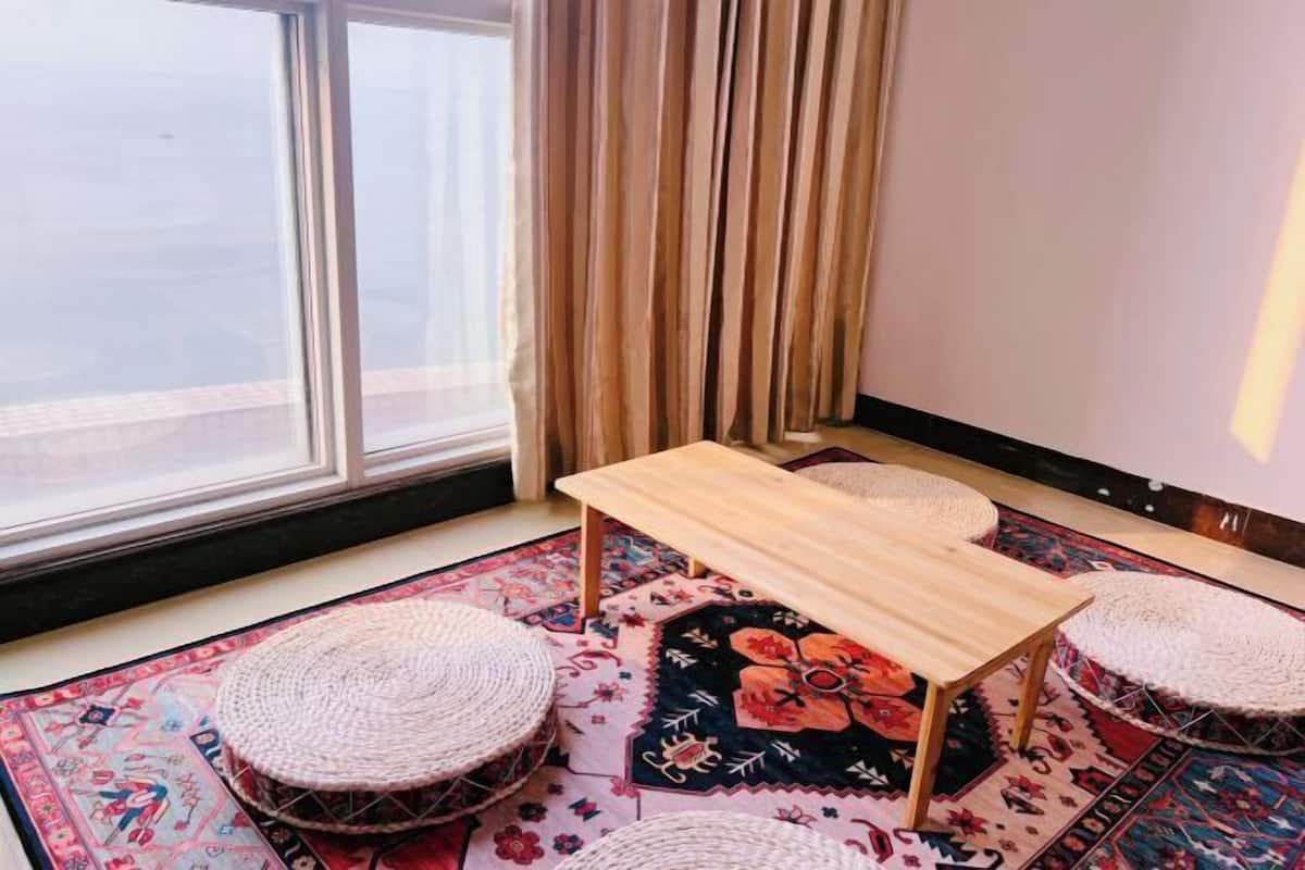 Una habitación con un ventanal grande, una mesa de madera, dos pufs tejidos y una alfombra estampada.