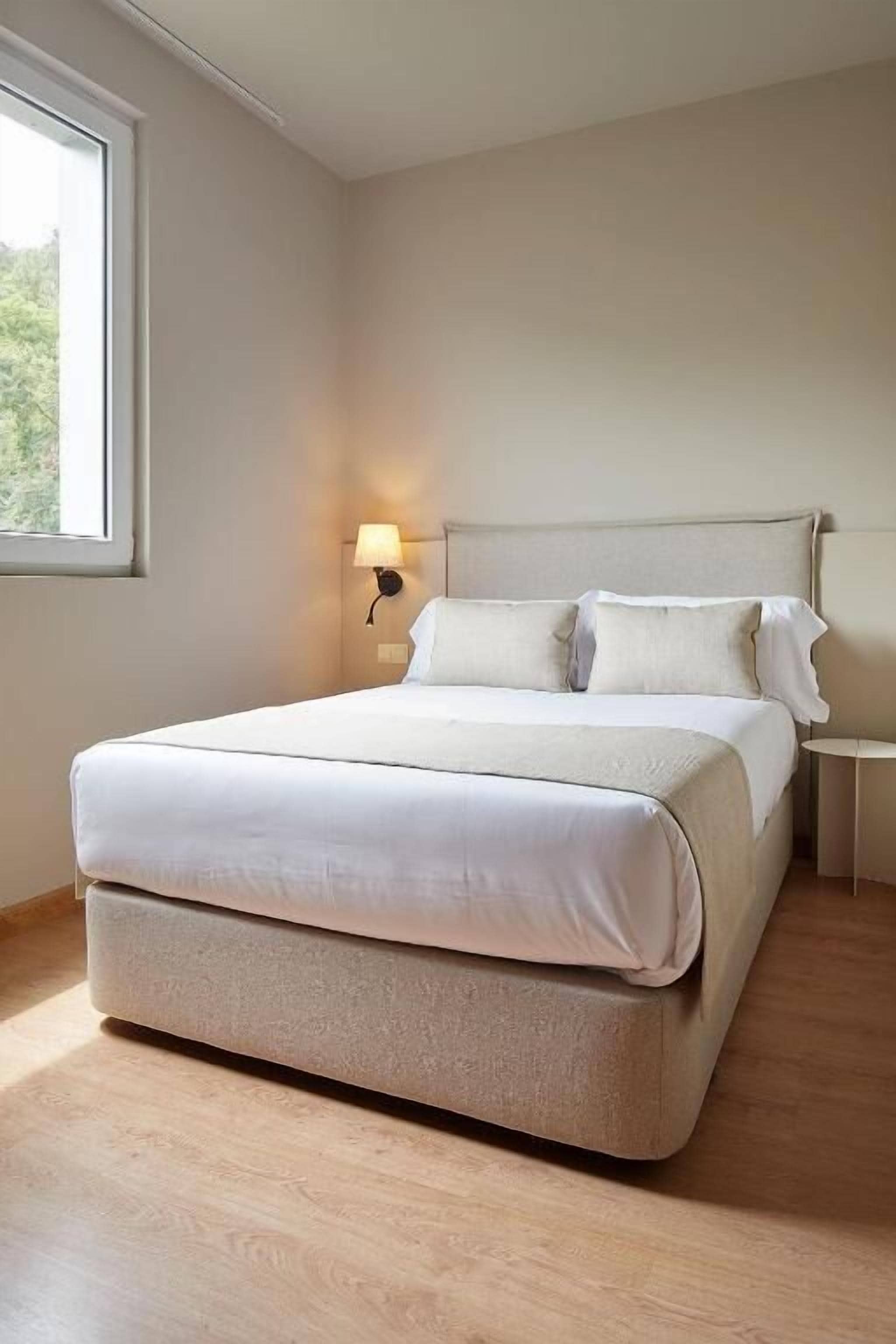 A neatly made bed in a room with a wooden floor, a nightstand with a lamp, and a window providing natural light.
