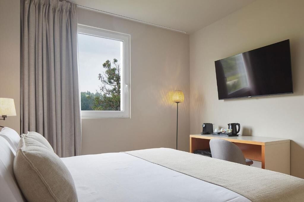 Una habitación de hotel con cama, escritorio, silla, televisión y ventana con cortinas.
