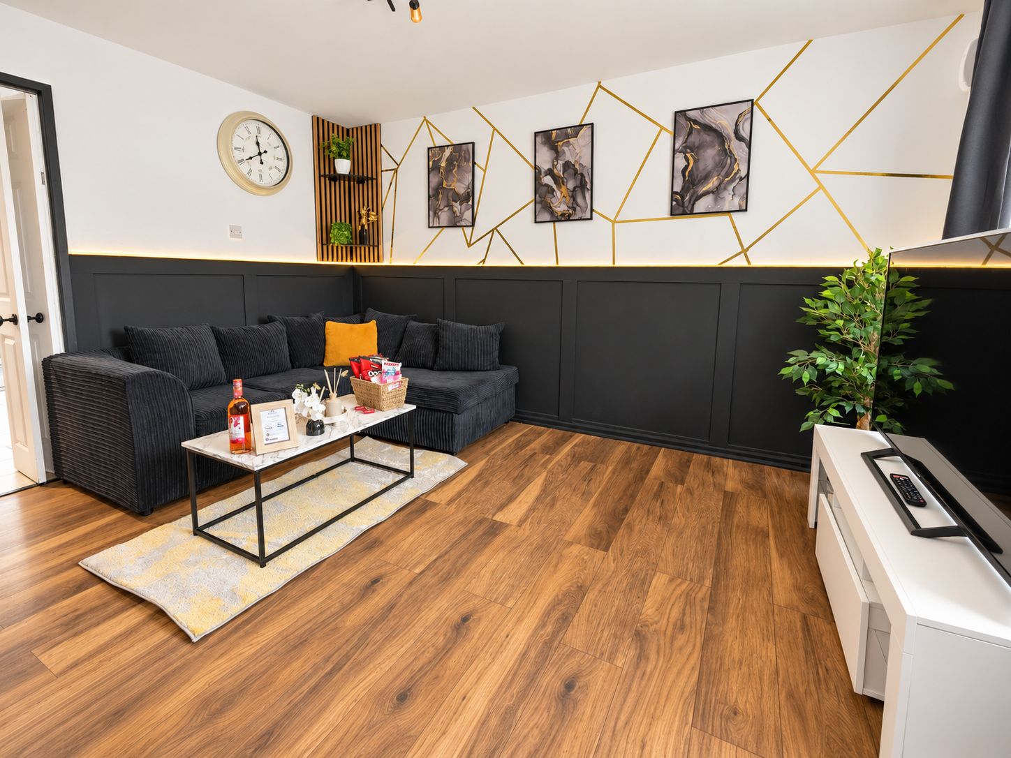 Un salon moderne comprenant un canapé d’angle, une table basse avec quelques objets décoratifs, une horloge murale, des œuvres d’art abstraites et un téléviseur.