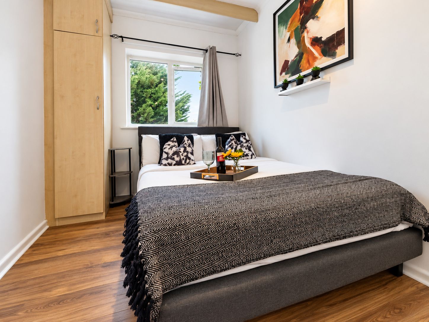A bedroom with a bed, a bedside table, a window with curtains, a painting on the wall, and a wooden wardrobe.