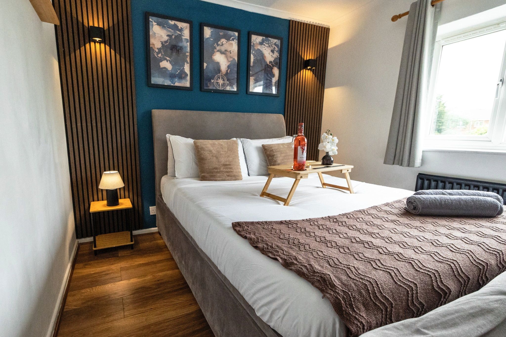 Ein modernes Schlafzimmer mit einem großen Bett, Nachttischlampen, einem kleinen Tisch mit einer Flasche und Kunstwerken an der Wand.