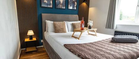 Een moderne slaapkamer met een groot bed, bedlampen, een klein tafeltje met een fles en kunst aan de muur.