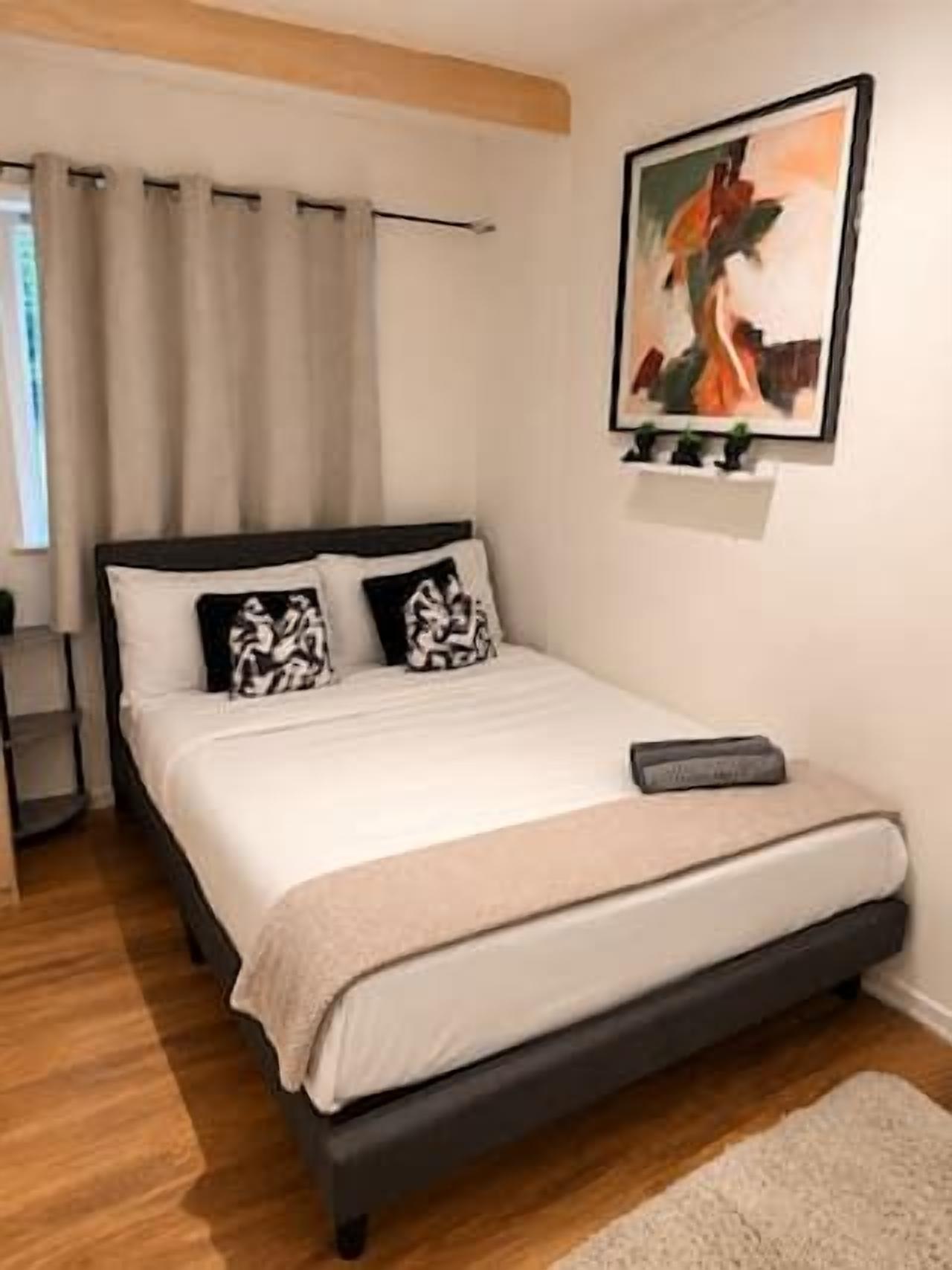 A bedroom with a bed, a nightstand, a painting, and a window with curtains.