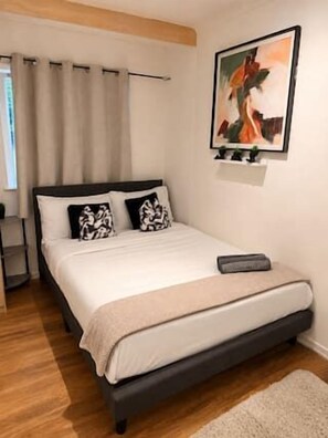 Quarto com cama, mesa de cabeceira, pintura e janela com cortinas.
