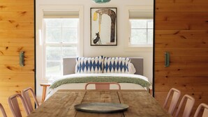 Een houten eettafel met stoelen, een hanglamp en een bed met een beddengoed met een patroon in een kamer met houten wanden en ramen.