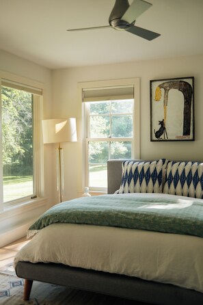 Un dormitorio con una cama, ventilador de techo, lámpara de pie y ventana con vista a árboles.