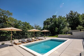 Une piscine rectangulaire, agrémentée de chaises longues et de parasols, entourée d’un jardin aménagé et d’un mur en pierre.