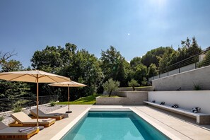 Un espace piscine extérieur moderne, doté de chaises longues et d’un parasol, entouré d’arbres et bordé d’un mur en béton.