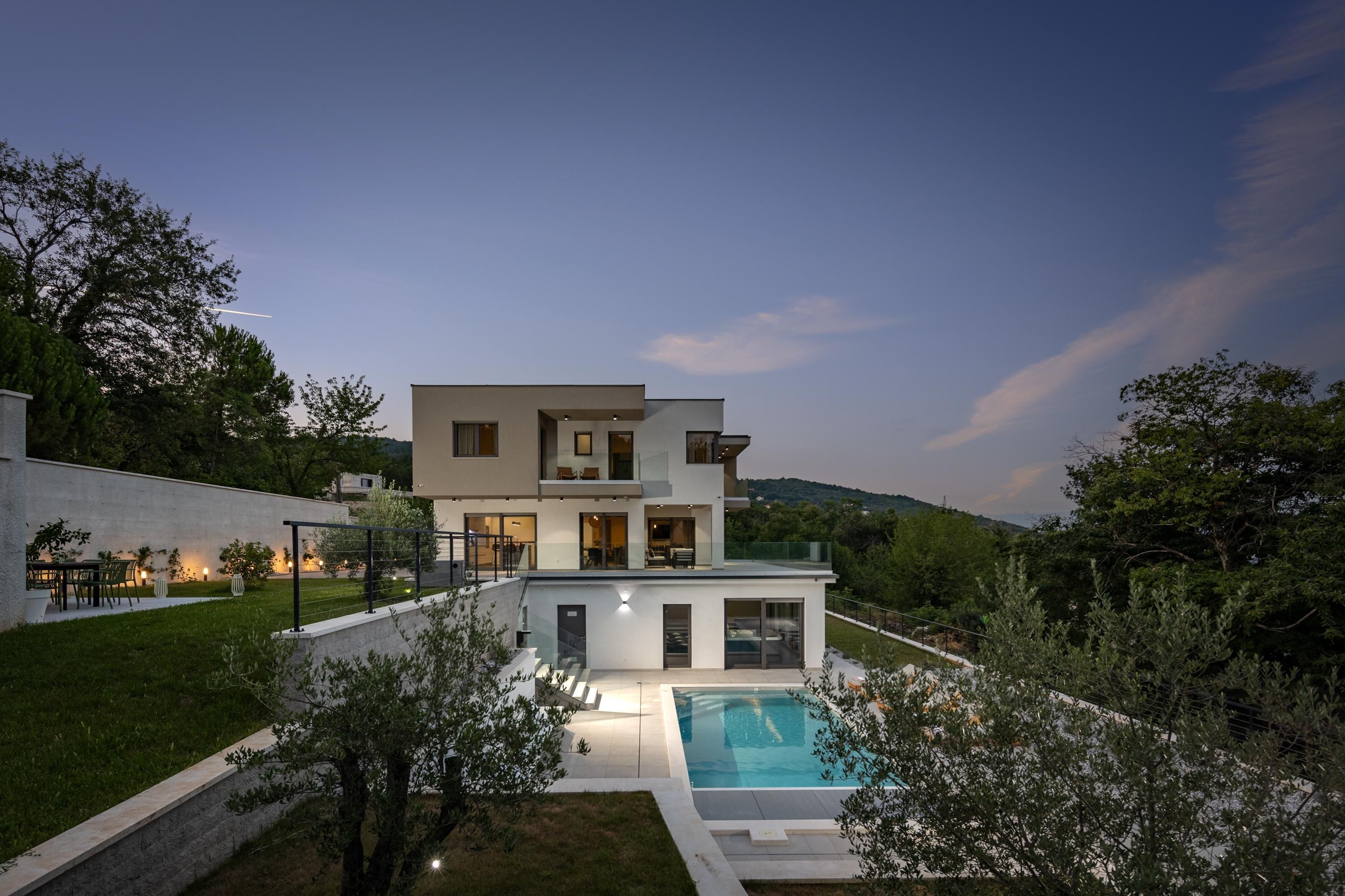 A modern two-story house with a swimming pool, surrounded by greenery and hills.