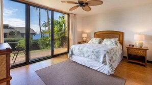 Un dormitorio con una cama, dos mesitas de noche con lámparas, un ventilador de techo y un amplio ventanal corredero que da a un balcón con vistas a palmeras y un edificio.