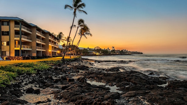 Un hotel frente a la playa con varios balcones que dan al océano, una costa rocosa y palmeras.
