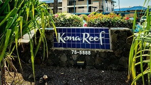 Letrero de Kona Reef con un muro de piedra, palmeras y un edificio con techos azules.