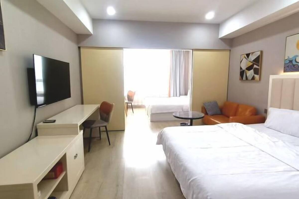 A hotel room with a bed, a desk with a TV, a chair, a sofa, and a window with curtains.