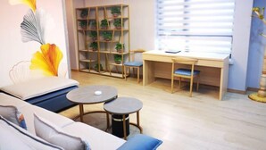 Ein Zimmer mit einer Couch, zwei kleinen Tischen, einem Holztisch mit Stühlen, einem B ücherregal mit Pflanzen und einem Fenster mit Jalousien.
