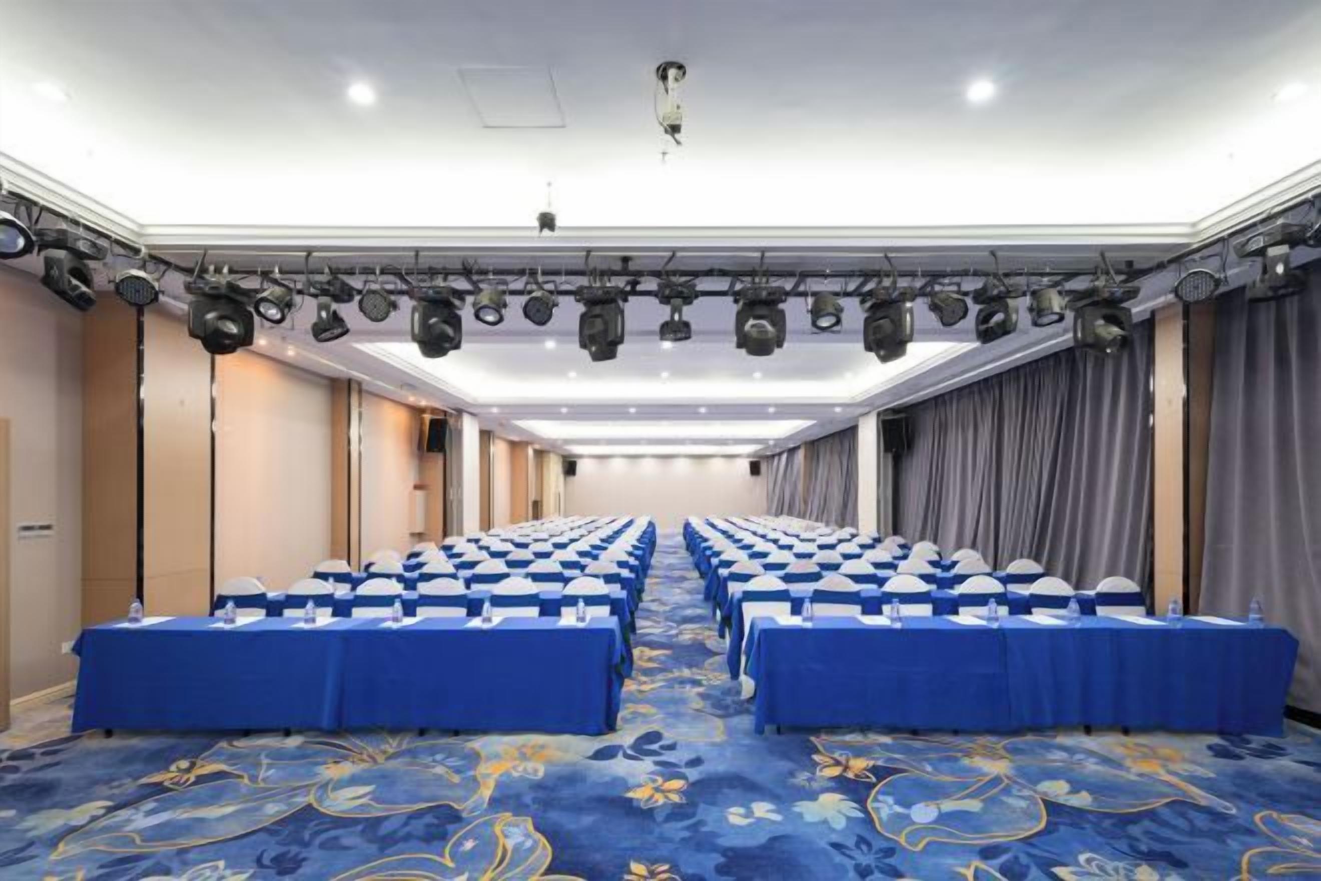 A conference room with rows of tables and chairs, a patterned carpet, and a ceiling with lighting fixtures.