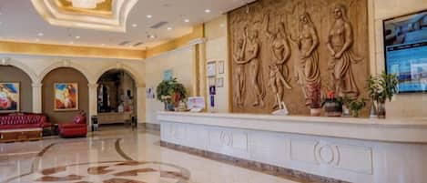 A hotel lobby with a reception desk, a large chandelier, and a wall with sculptural reliefs.