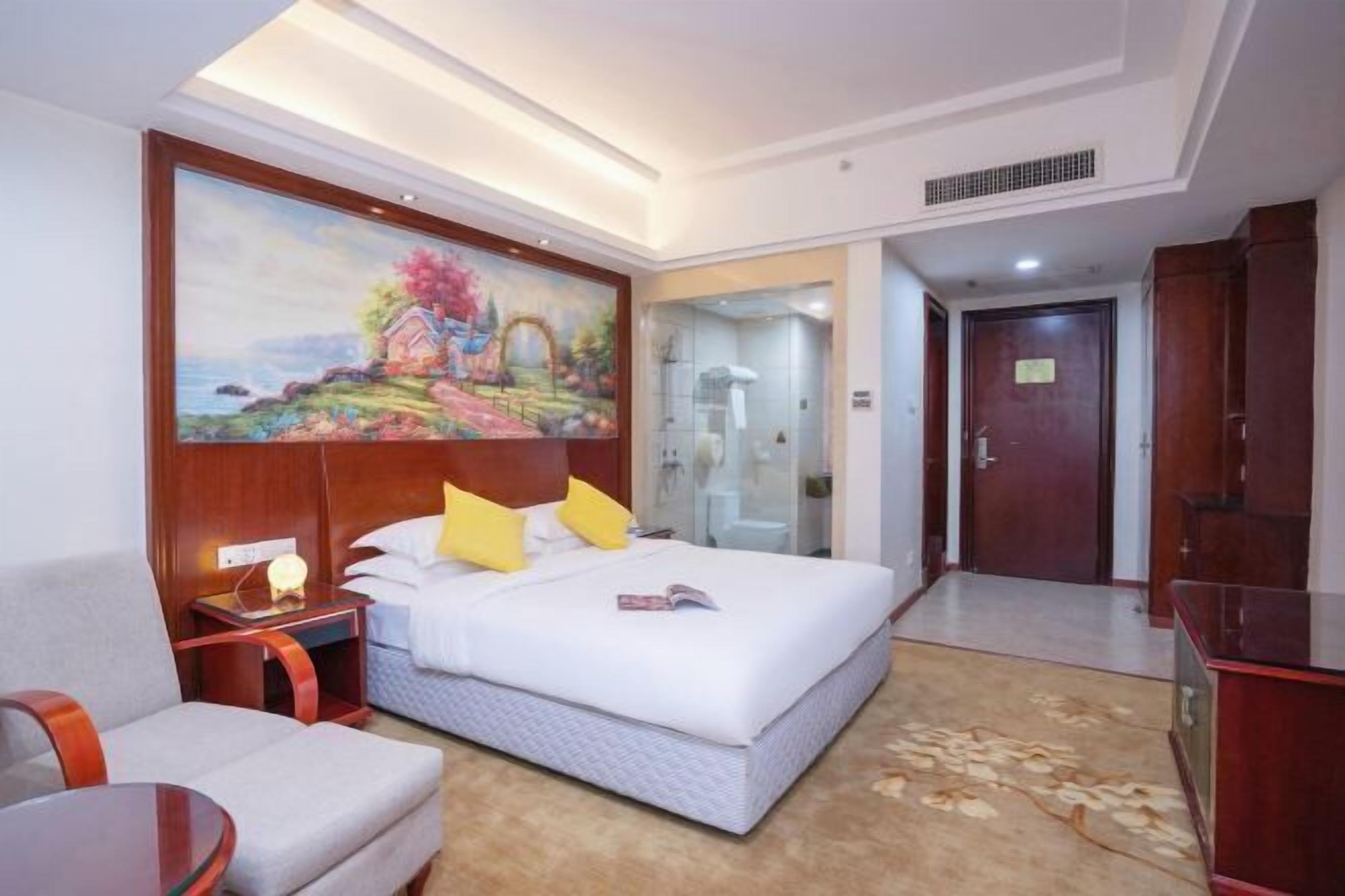 Ein Hotelzimmer mit einem großen Bett, einem Bild an der Wand, einem Schreibtisch, einem Sessel und einem durch eine Glastür einsehbaren Badezimmer.