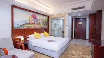 Quarto de hotel com uma cama grande, um quadro na parede, uma escrivaninha, uma cadeira e um banheiro visível através de uma porta de vidro.