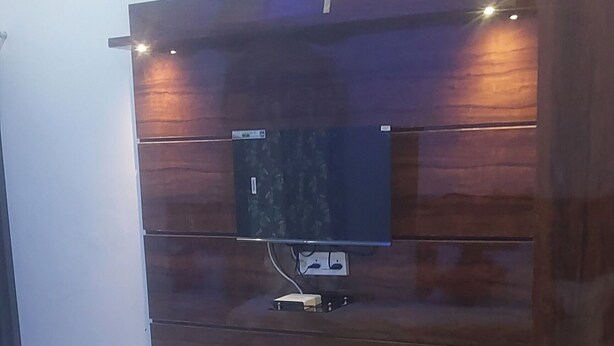 A modern bedroom with a bed, a TV mounted on a wooden wall, and a small shelf. - Osho cottage  (Rajnagar)