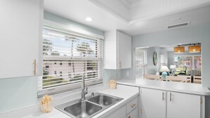 A modern kitchen with white cabinetry, a stainless steel sink, and a window with blinds. - Beachy Butterfly! Poolside Holmes Beach Stay! (Holmes Beach)