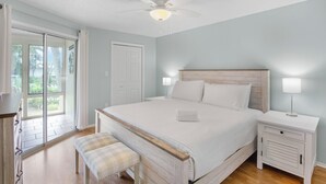 A bedroom with a large bed, a nightstand, a bench, a ceiling fan, and a sliding glass door leading to a balcony. - Beachy Butterfly! Poolside Holmes Beach Stay! (Holmes Beach)