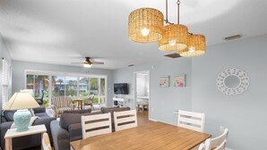A modern living room with a wooden dining table, white chairs, a sofa, and a television. - Beachy Butterfly! Poolside Holmes Beach Stay! (Holmes Beach)