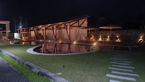 Een zwembad buiten met verlichting, een houten pergola, lounge stoelen en parasols.
