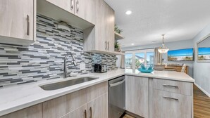 Una cocina moderna con una isla grande, salpicadero de azulejos y vista a la playa a través de amplias ventanas.