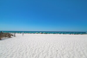 Ein sandiger Strand mit einer Holzlattenzaun, türkisfarbenen Sonnenschirmen und dem Meer im Hintergrund.