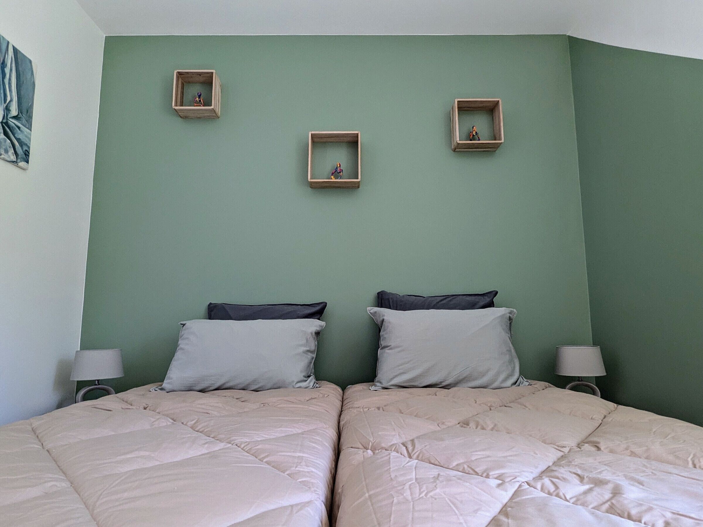 Une chambre avec deux lits, un mur vert et trois petites étagères en bois ornées de figurines.