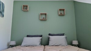 Une chambre avec deux lits, un mur vert et trois petites étagères en bois ornées de figurines.