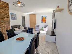 A modern living room with a dining area, a fireplace, and a TV mounted on the wall. - Single-storey house with enclosed garden in Longchamps (Longchamps-sur-Aire)