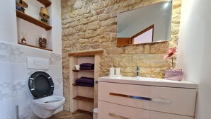 A bathroom with a stone wall, a white toilet, a vanity with drawers, and shelves with towels and toiletries. - Comfortable room with private bathroom (Pierrepont)