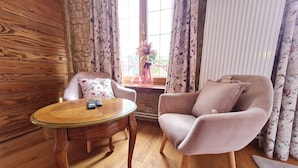 A cozy room with a wooden floor, a round table with two remotes, a plush armchair, and a window with floral curtains. - Comfortable room with private bathroom (Pierrepont)