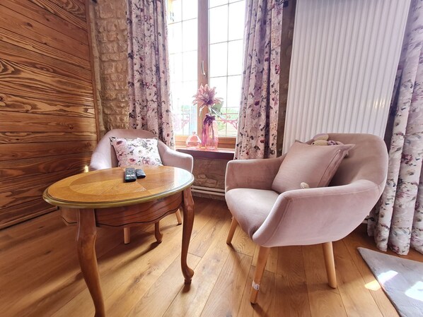 A cozy room with a wooden floor, a round table with two remotes, a plush armchair, and a window with floral curtains. - Comfortable room with private bathroom (Pierrepont)