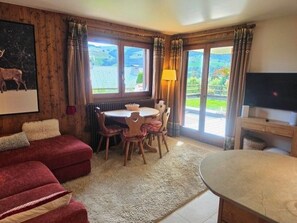 A room with a red sofa, a dining table with chairs, a television, and large windows with curtains. - Cozy apartment with terrace in Megève, perfect for 2-3 guests (Megève)