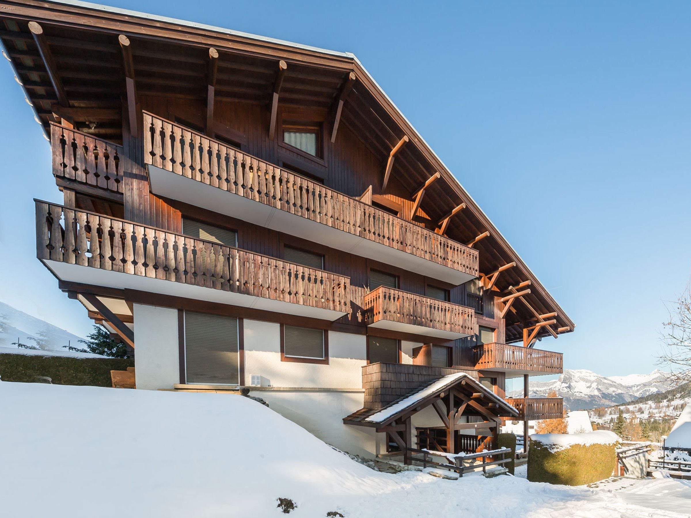 A multi-story wooden chalet with balconies, set against a snowy mountain backdrop.