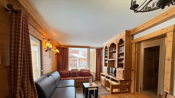 Une pièce chaleureuse avec un banc en bois, un canapé et une table en bois. On y trouve une fenêtre avec des rideaux, une bibliothèque et une porte.