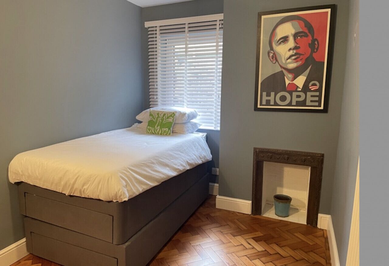 Une chambre à coucher avec un lit, une fenêtre avec des stores, une cheminée et une affiche accrochée au mur.