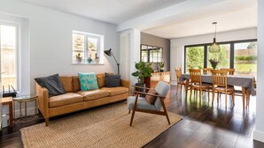 Un salon moderne avec un canapé en cuir, un coin repas avec des chaises et une table en bois, et de grandes fenêtres offrant une vue sur l’extérieur.