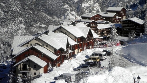 A snow-covered mountain village with wooden buildings, a ski slope, and a few parked vehicles.