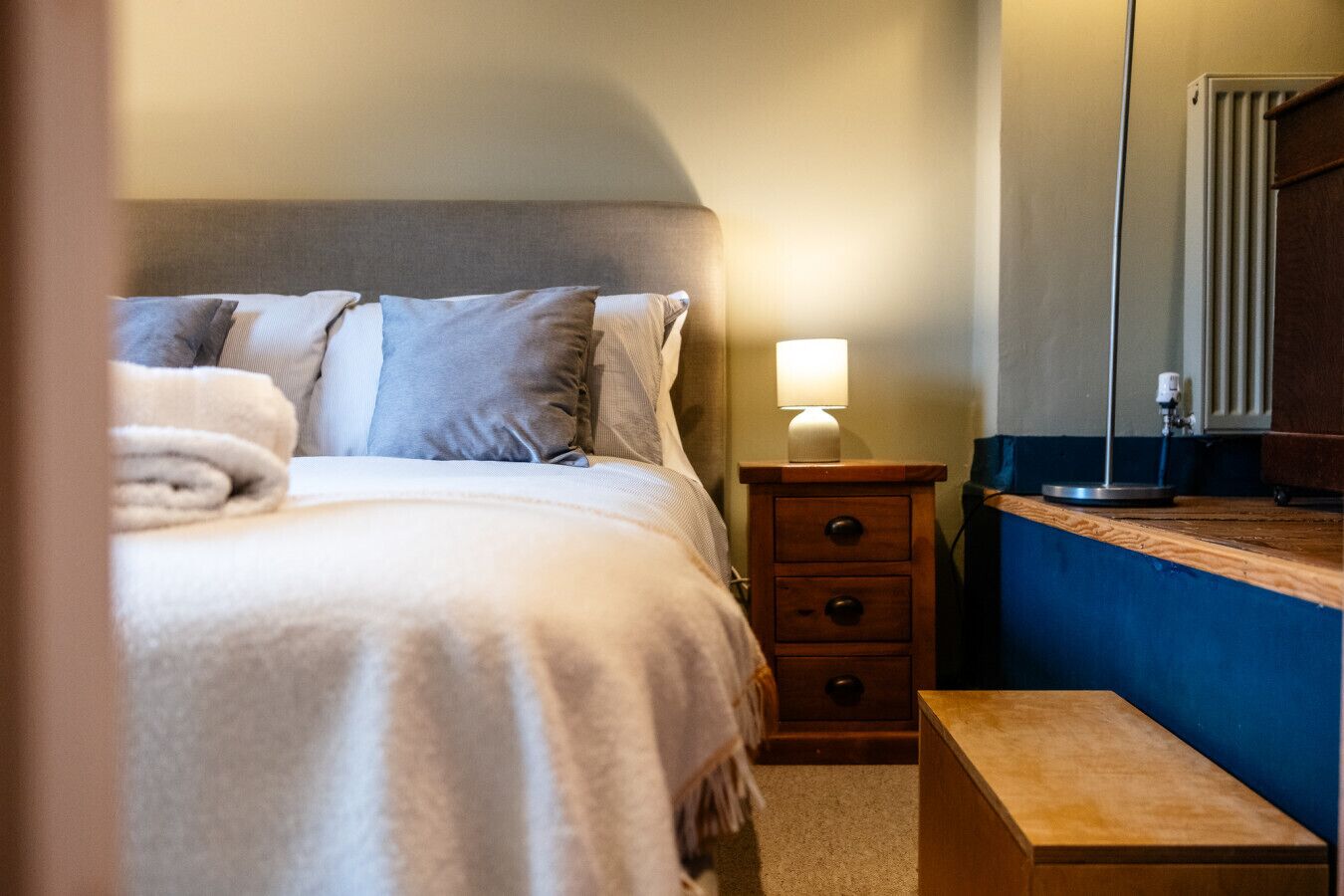 Una cama bien hecha con almohadas, una lámpara de noche, una mesita de noche de madera y un pequeño banco de madera.