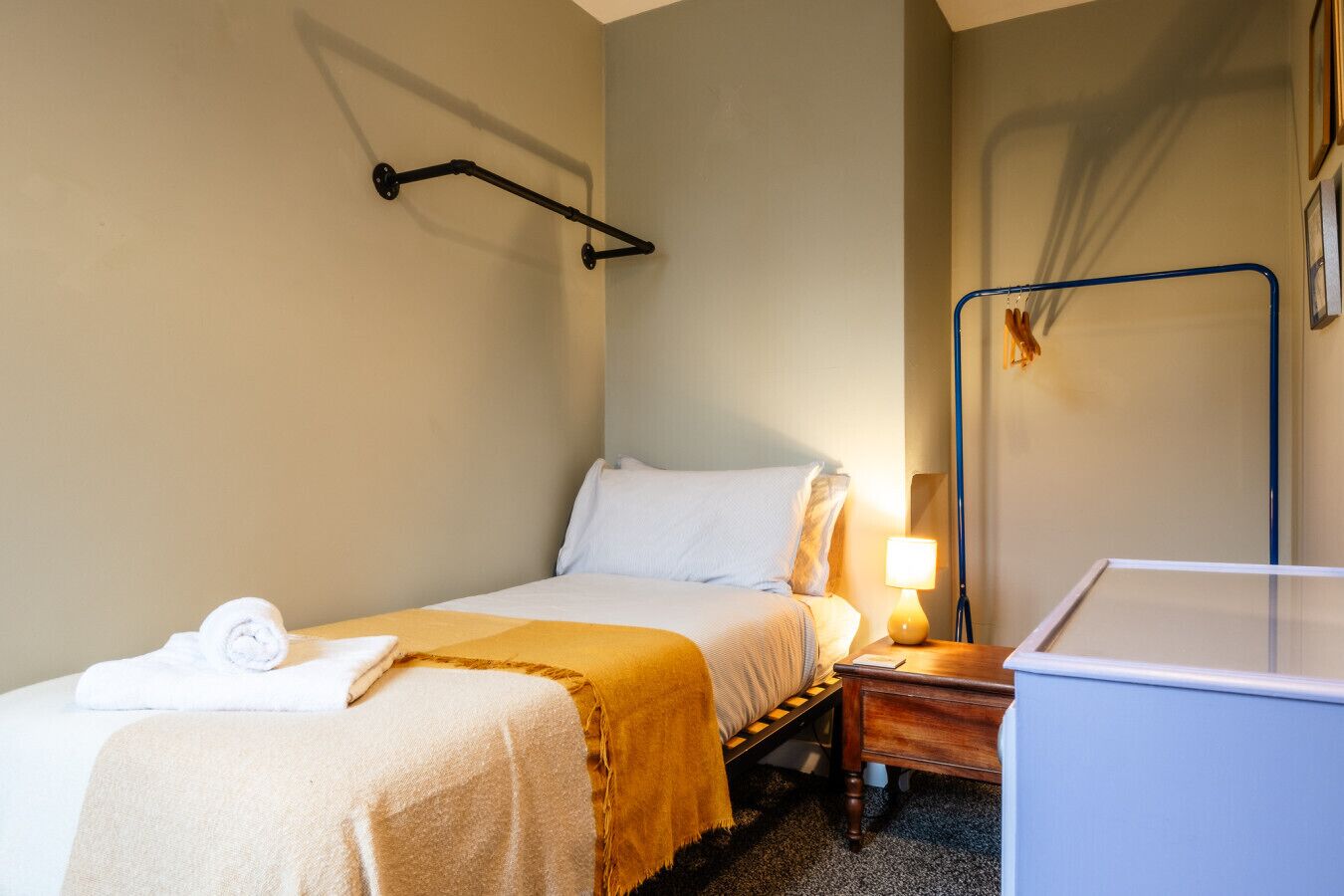 Una cama individual con un cojín blanco y una toalla enrollada, una mesita de noche con una lámpara y un escritorio azul en una habitación de hotel.