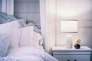 A neatly made bed with white linens, a white nightstand with a lamp and a vase of flowers, and a blue upholstered headboard.